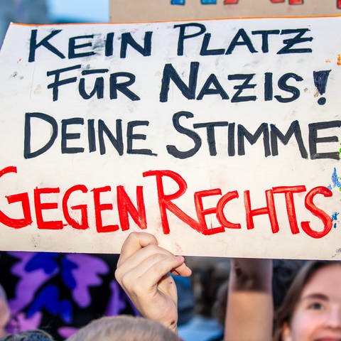 Auf einer Demo wird ein Plakat mit der Aufschrift: "Kein Platz für Nazis! Deine Stimme gegen Rechts" hochgehalten.
