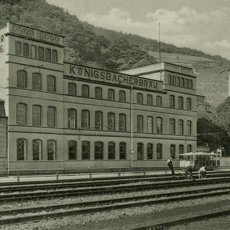 Brauereigebäude mit Biergarten, im Vordergrund Eisenbahngleise, vor der Brauerei Straßenbahnwagen, im Hintergrund rechts Weinberge; um 1930 (Foto: Stadtarchiv Koblenz)