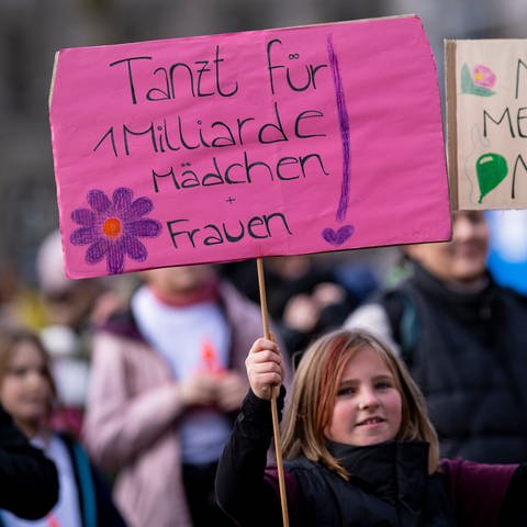 Eine Teilnehmerin eines Tanz-Flashmobs und einer Demonstration gegen Gewalt an Frauen und Mädchen hält zwei Plakate mit der Aufschrift «Tanzt für 1 Milliarde Mädchen und Frauen» und «No means No!».