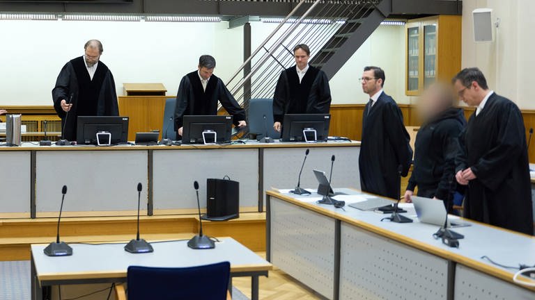 Das Gericht, der Angeklagte und seine Verteidiger stehen im Gerichtssaal des Oberlandesgerichts Koblenz. 