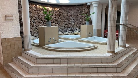 Auf einem erhöhten Bereich sind vier kleinere, weiße Becken in den Boden eingelassen. Die Wand des Bereichs ist mit braunem Naturstein verkleidet. (Foto: SWR)