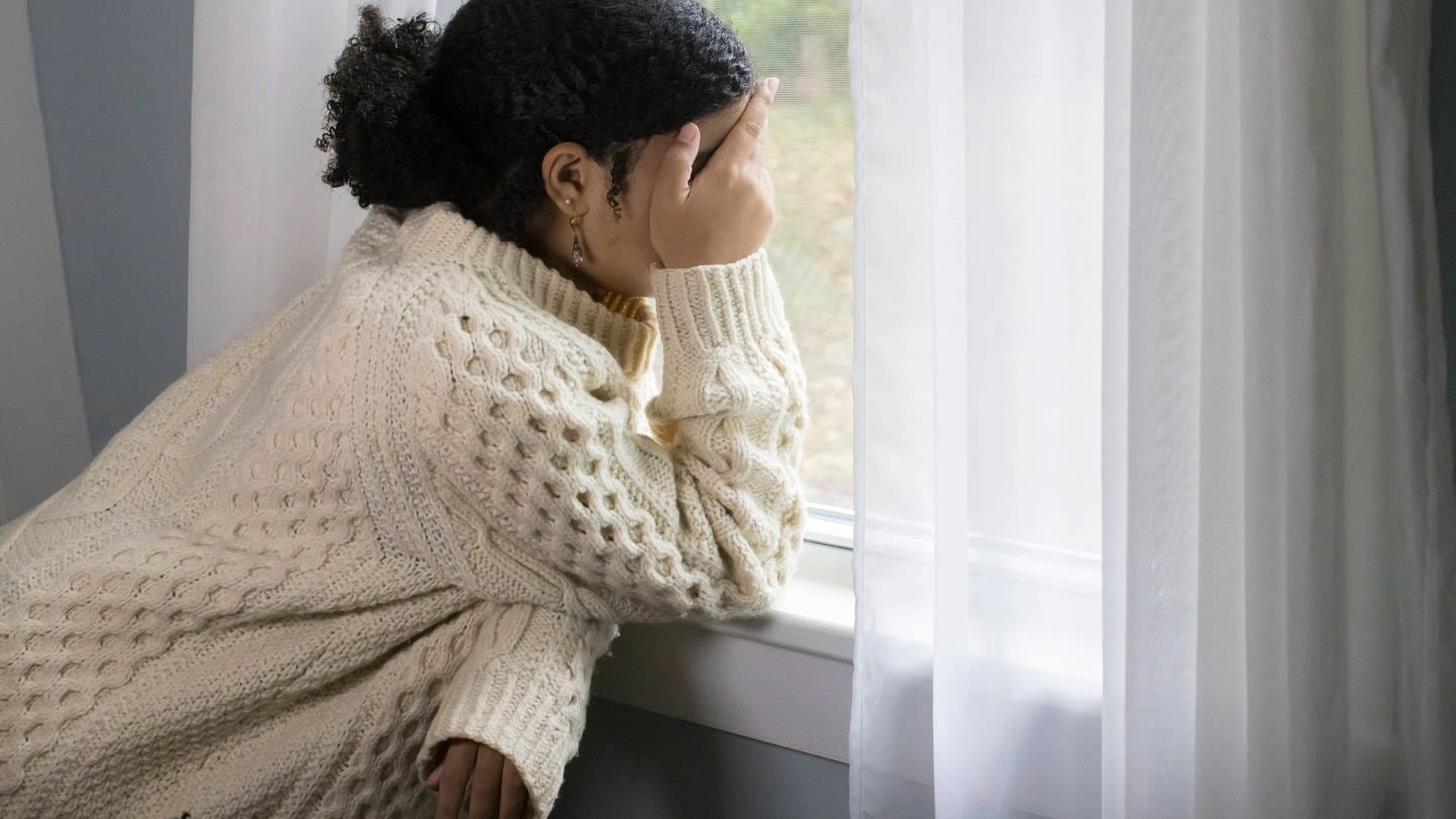 Junge Frau lehnt am Fenster mit der Hand vor dem Gesicht - mit einer neuen Strategie will die Bundesregierung die Zahl der Suizide senken. (Foto: IMAGO, IMAGO / Cavan Images)