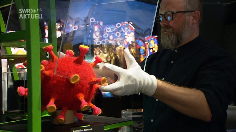 Museumsmitarbeiter stülpt Glasvitrine über Corona-Virus, nachgestellt aus Wolle (Foto: SWR)
