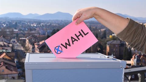 Stilisierter Wahlzettel wird in eine Box eingeworfen und im Hintergrund ist verschwommen die Stadt Landau zu erkennen (Foto: Stadt Landau)