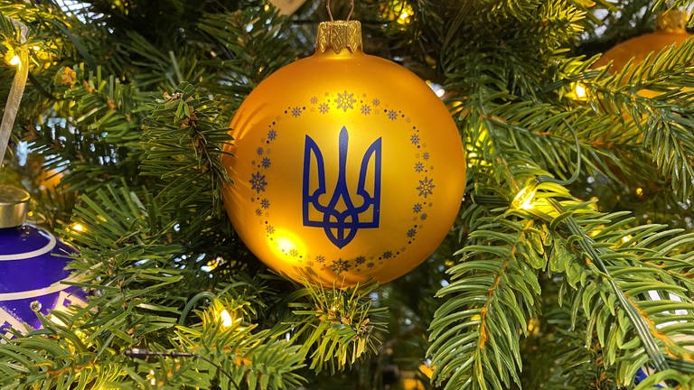 An einem geschmückten Baum in einem Geschäft hängt eine Kugel in den ukrainischen Nationalfarben Blau und Gelb mit dem Dreizack als Wappen. (Foto: dpa Bildfunk, picture alliance/dpa | Ulf Mauder)