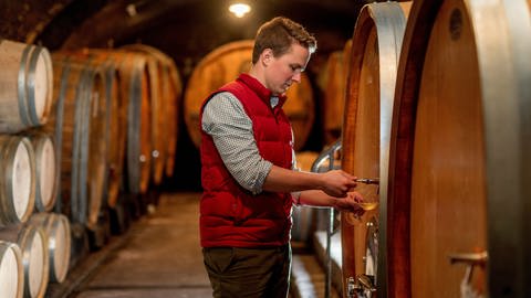 Hans-Christoph Stolleis zapft Wein aus einem Weinfass in seinem Weinkeller. Er trägt ein kariertes Hemd und eine rote Weste. (Foto: Privat)
