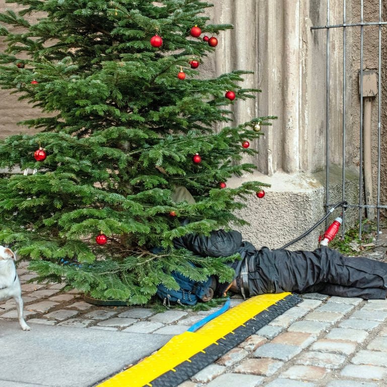 Obdachloser liegt unter einem geschmückten Tannenbaum offenbar an einer Weihnachtsmarkt-Bude Symbolbild für Hilfe für Obdachlose in eiskalten Nächten (Foto: IMAGO, Wolfgang Maria Weber)