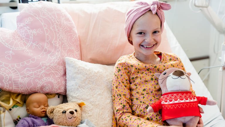 Emilia sitzt lächelns auf einem Krankenbett, in dem rosafarbene Kissen. Sie hält ein Plüschtier in den Händen und hat ein über der Stirn geknotetes Kopftuch auf.  (Foto: Kinderkrebshilfe Mainz)