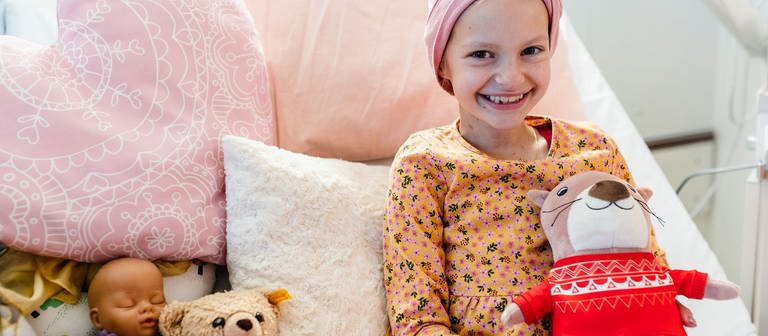 Emilia sitzt lächelns auf einem Krankenbett, in dem rosafarbene Kissen. Sie hält ein Plüschtier in den Händen und hat ein über der Stirn geknotetes Kopftuch auf.  (Foto: Kinderkrebshilfe Mainz)
