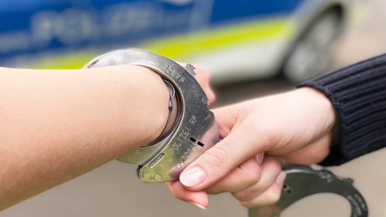 Ein Polizist hat einem Menschen Handschellen angelegt. Im Hintergrund ist ein Polizeiauto zu sehen.  (Foto: SWR, D. Brusch (Symbolbild))