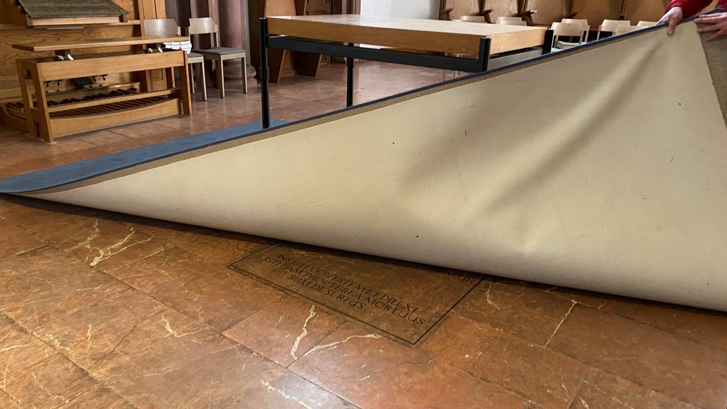 Im Altarraum der Kirche wird ein Teppich angehoben, darunter ist eine Grabplatte. (Foto: SWR)