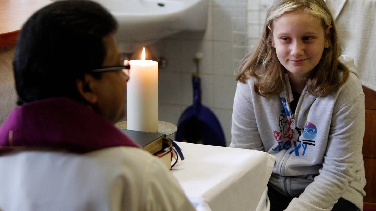 Ein Mädchen sitzt mit Pfarrer in einem Raum. Ihm wird die Erstbeichte abgenommen. (Foto: IMAGO, IMAGO / Funke Foto Services)
