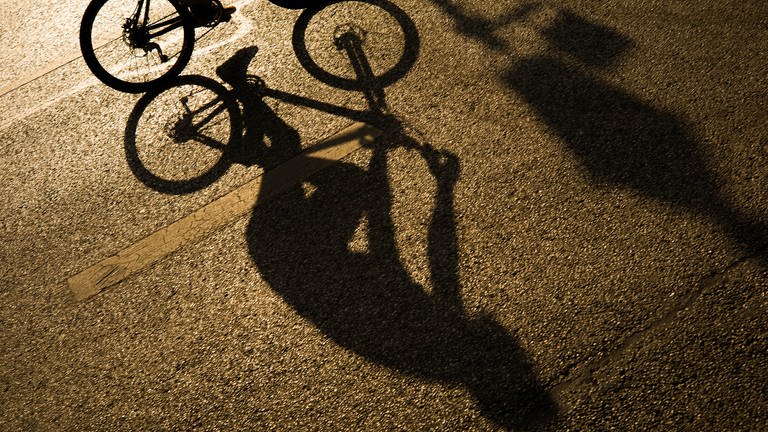 picture alliancedpa | Peter Kneffel (Foto: dpa Bildfunk, Der Schatten eines Radfahrers zeichnet sich im Licht der Abendsonne auf dem Asphalt einer Straße ab. Dooring-Unfälle stellen in Mainz eine Gefahr für Radfahrer da.)