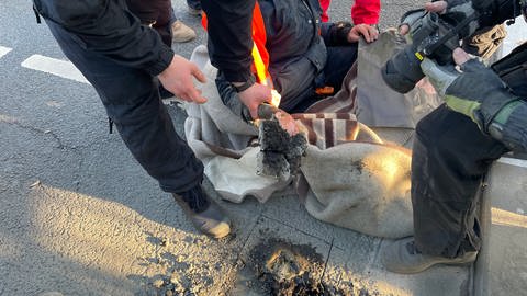 Einer der Klimaaktivisten hatte sich so fest an der Straße festgeklebt, dass seine Hand mithilfe einer Flex zusammen mit einem Stück Asphalt aus der Straße geschnitten werden musste.