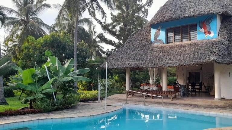 Das Haus, das die Gemeinde Wöllstein in Kenia geerbt hat, hat ein Reetdach. Im Giebel an der Hauswand wurden zwei Greifvögel aufgemalt. Vor dem Haus gibt es einen Pool. 