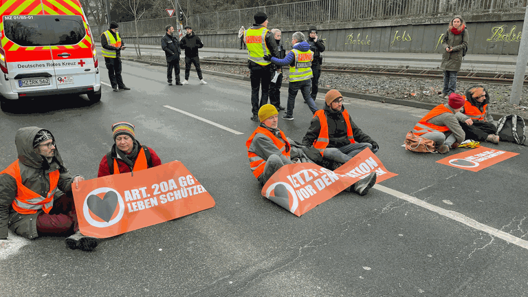 Klimaaktivisten der Letzten Generation haben am Freitagmorgen die Saarstraße in Mainz blockiert. Auf der Straße saßen sechs Menschen. Wegen der kalten Temperaturenhaben hatten sie sich warm angezogen. Einige haben sich an der Straße festgeklebt. Es gab lange Staus. 