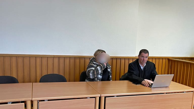 Eine Angeklagte sitzt mit Maske im Amtsgericht in Bad Sobernheim neben ihrem Anwalt. Sie hat blonde Strähnchen und längere Haare und trägt ein schwarzes T-Shirt und eine karierte Hemd-Jacke. (Foto: SWR, I. Hartmann)