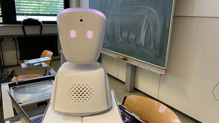 Ein kleiner Roboter steht auf dem Lehrerpult in einem Klassenzimmer vor einer Tafel.