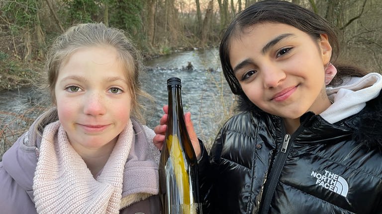 Milla und Mina stehen am Guldenbach und halten ihre Flaschenpost in den Händen.