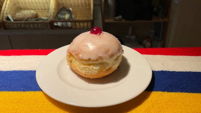 Ein Eierlikörkreppel mit Zuckerguss und einem kleinen Klecks Marmelade liegt auf einem Teller, der auf einem bunten Fastnachtsschal liegt. (Foto: SWR, Alexander Dietz)