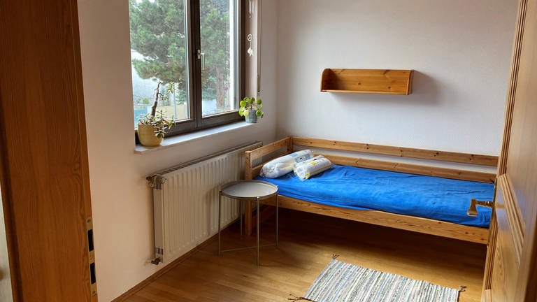 Blick in ein Zimmer mit Bett, Nachttisch und einem kleinen Regal an der Wand.