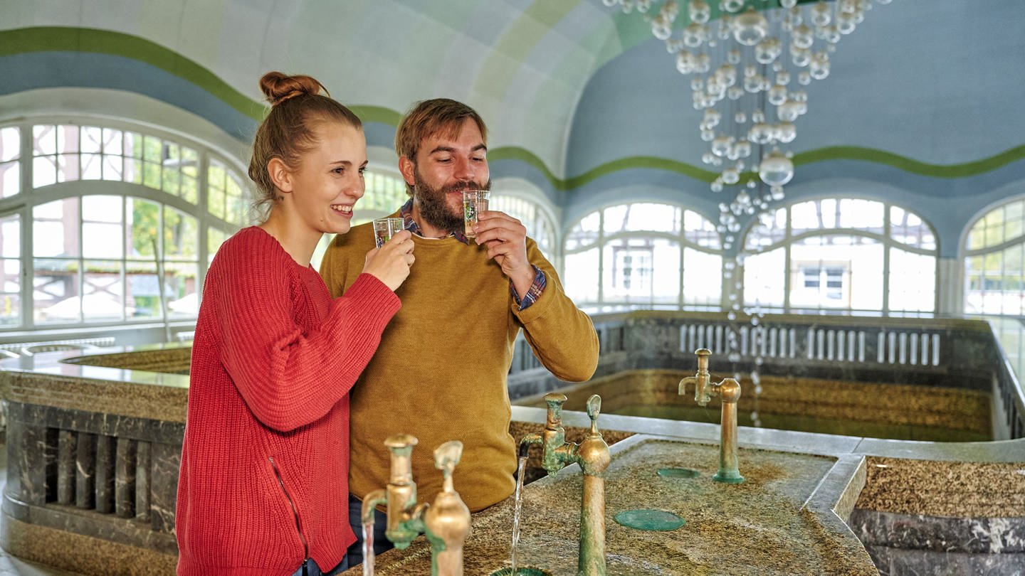 Ein Paar trinkt Solewasser aus den Hähnen in der Brunnenhalle in Bad Münster am Stein (Foto: Rheinland-Pfalz-Touristik GmbH, Florian Trykowski)