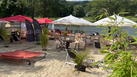 Stühle und Tische stehen unter Sonnenschirmen und Palmen im Sandstrand. Die Beachbar in Minden lädt nach dem Hochwasser 2021 wieder zum erholen ein. (Foto: SWR, Jana Hausmann)