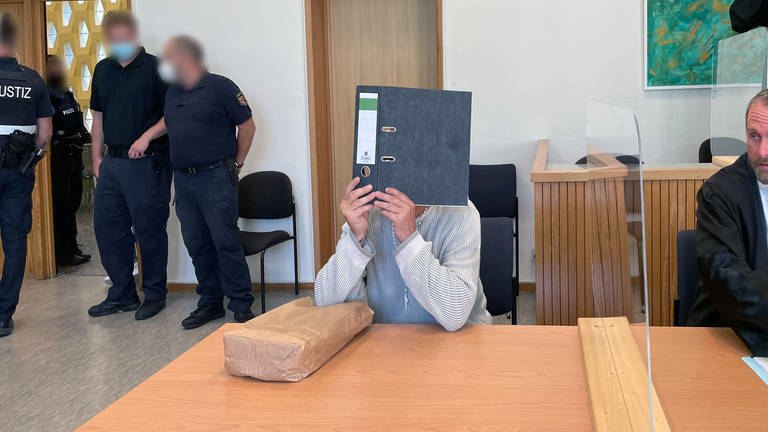 Der Angeklagter sitzt im Gerichtssaal beim Prozess in Idar-Oberstein. Er verdeckt mit einem Aktenordner sein Gesicht. Dem 55-Jährigen wird vorgeworfen, im Februar 2022 im Internet dazu aufgerufen zu haben, Polizisten zu erschießen. (Foto: SWR)