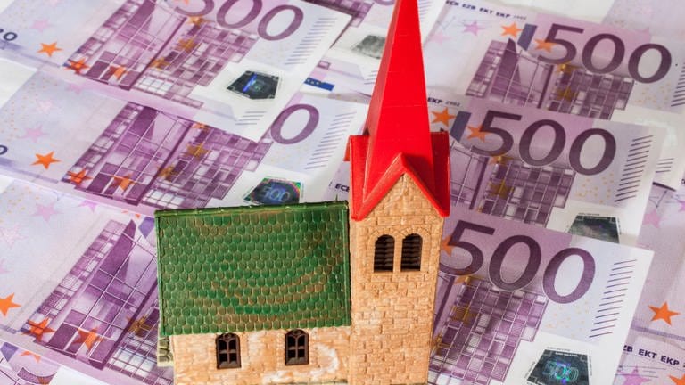 Eine Spielzeugkirche steht auf einem Untergrund auf 500-Euro-Scheinen. Geld wird im Bistum Trier knapp. Es muss Millionen Euro einsparen. (Foto: IMAGO, Jochen Eckel )