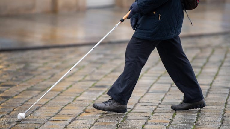 Eine blinde Person geht mit ihrem Blindenstock eine Straße entlang.