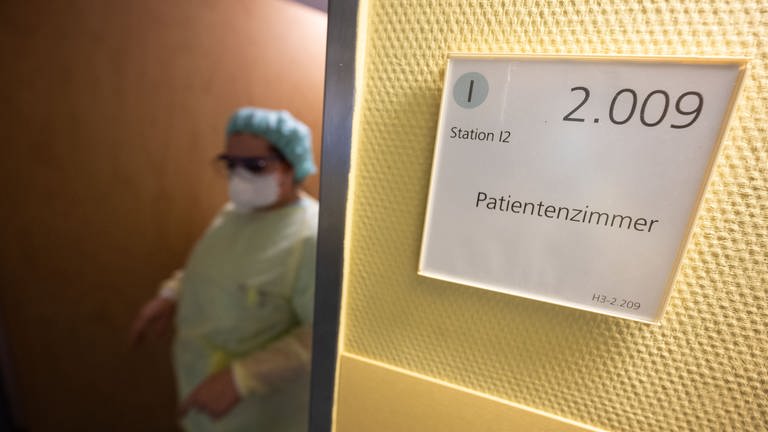 Eine Krankenpflegerin verlässt in Schutzkleidung ein Patientenzimmer. Es ist ein neues Phänomen: Die Corona-Pandemie verlagert sich stärker auf die Normalstationen der Krankenhäuser. 