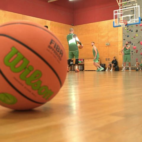 Basketball liegt auf dem Boden einer Sporthalle, im Hintergrund Basketballspieler und ein Korb