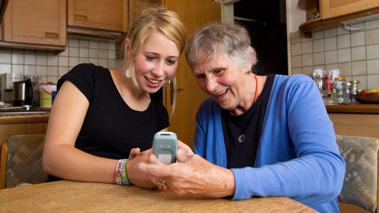 Pflegerin zeigt im Rahmen der ambulanten Pflege älterer Frau, wie ein Mobiltelefon funktioniert.
