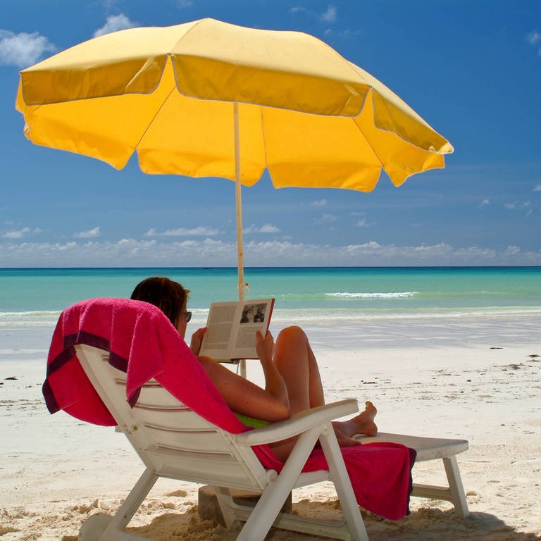 Eine Frau liest ein Buch unter einem Sonnenschirm am Strand (Foto: IMAGO, imagebroker)