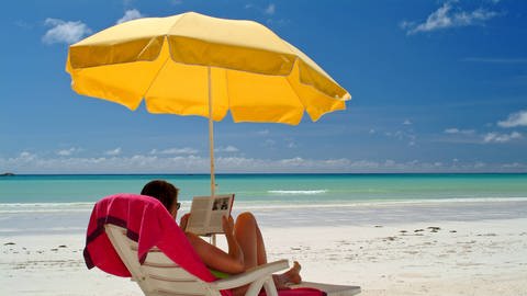 Eine Frau liest ein Buch unter einem Sonnenschirm am Strand (Foto: IMAGO, imagebroker)