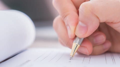 Eine Hand streicht mit einem Kugelschreiber Textzeilen auf einem Blatt.