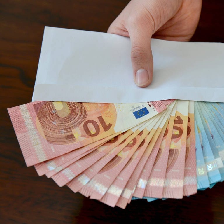 Eine Hand hält einen Umschlag, aus dem gefächerte Euroscheine herausschauen. (Foto: IMAGO, Joko)
