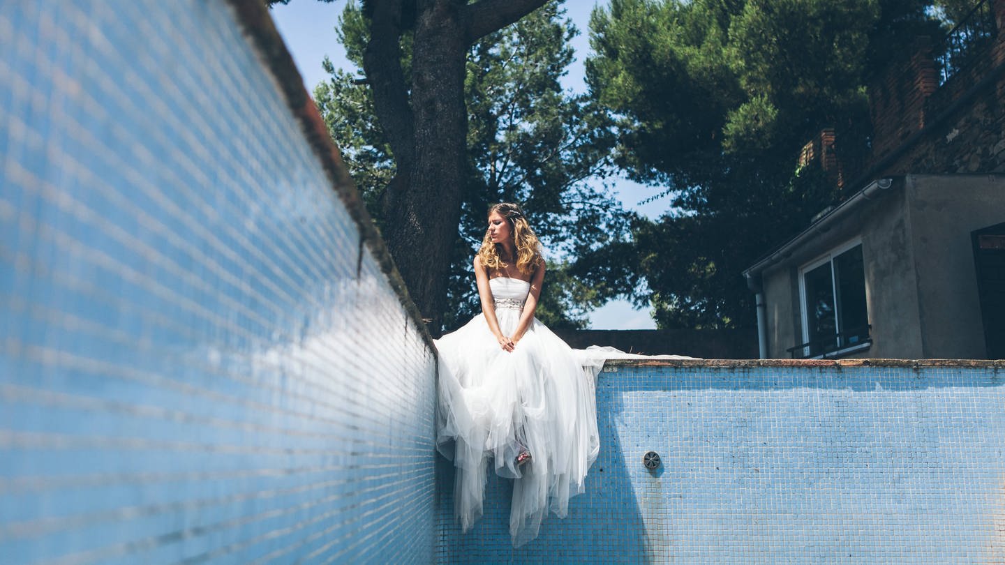 Eine junge Frau im weißen Brautkleid sitzt am Rand eines leeren Schwimmbeckens und sieht mäßig glücklich aus (Foto: IMAGO, IMAGO / Addictive Stock)