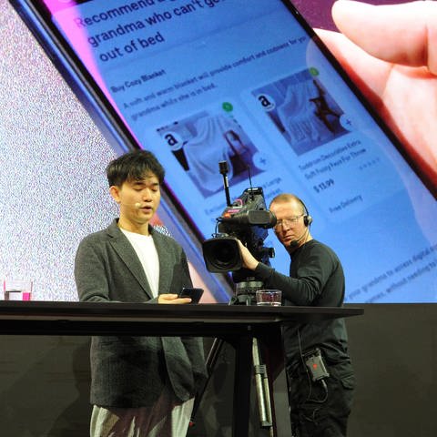 Jerry Yue, Gründer und Chef des US-Technologieunternehmens Brain Technologies, steht beim Mobile World Congress (MWC) auf einer Bühne und präsentiert einen Prototypen eines Künstliche-Intelligenz-Smartphones der Deutschen Telekom. Auf der Leinwand hinter ihm sieht man Geschenkvorschläge, welche die KI Yue auf dem Handydisplay gemacht hat