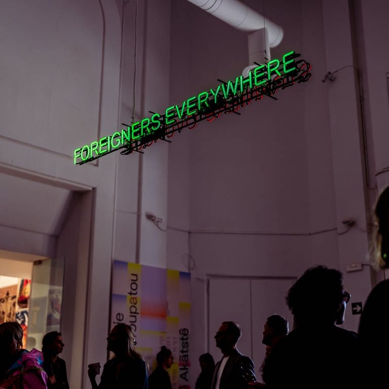 Besucher in einem dunklen Raum mit der Neon-Schrift: "Foreigners Everywhere" (Foto: La Biennale di Venezia)