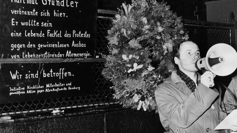 Pastor Ulfried Kleinert hält vor der Gedenktafel eine Rede. Am 22.11.1977 wurde vor der Petri-Kirche in Hamburg an der Stelle, an der sich der Tübinger Lehrer Hartmut Gründler aus Protest gegen die Kernenergie verbrannt hatte, eine Gedenktafel enthüllt. (Foto: dpa Bildfunk, picture-alliance/ dpa | Lothar Heidtmann)