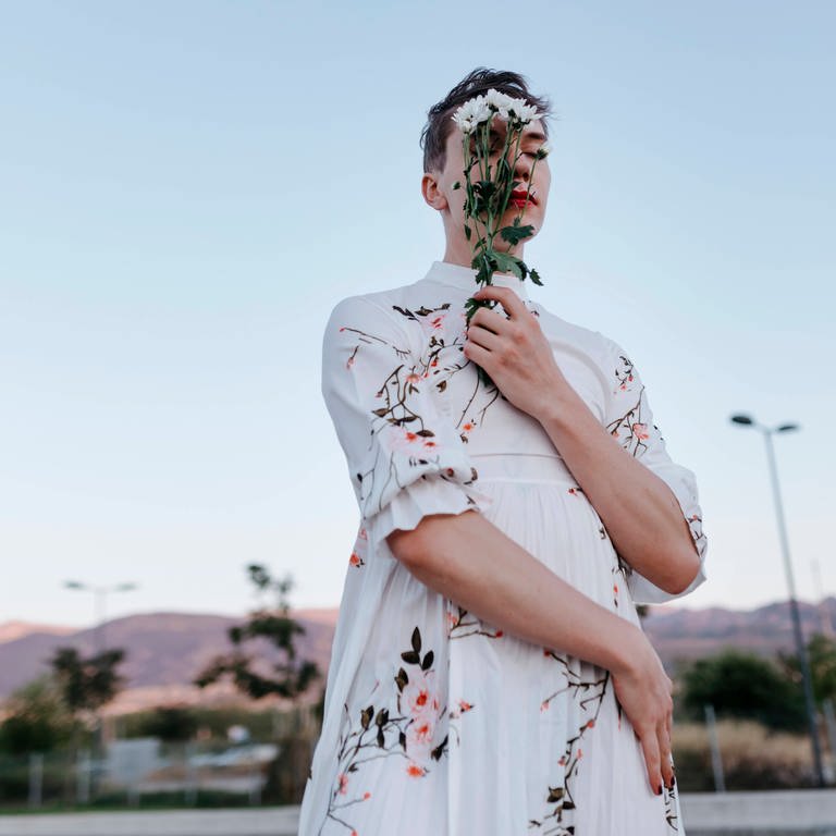 Gender fluid man covering face with bouquet of daisies while standing on street during sunset.