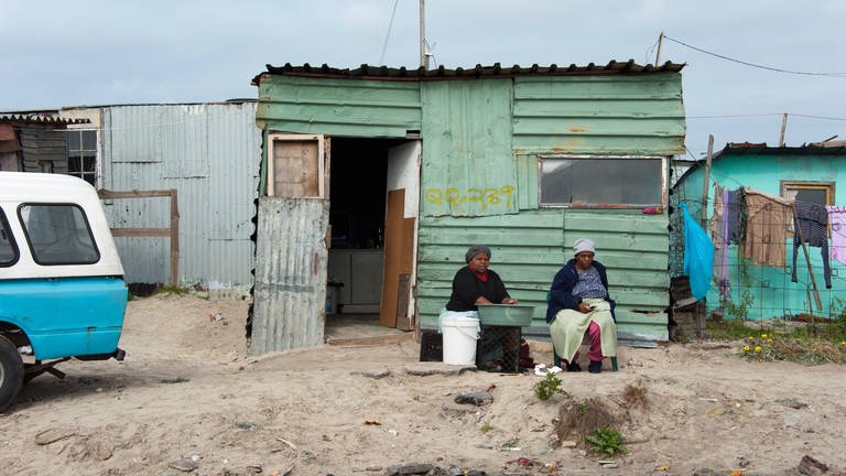 Zwei Frauen sitzen vor einer Wellblechhütte im einem Armenviertel bei Kapstadt, Südafrika (Foto: IMAGO, imago images / blickwinkel/McPhoto/ Frauke Scholz)
