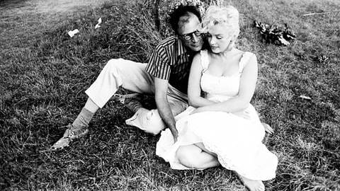Marilyn Monroes Ehemann, der Dramatiker Arthur Miller schrieb für sie unter anderem das Drehbuch zu „The Misfits — Nicht gesellschaftsfähig“: Schwarz-weiß Bild eines Paares, das umschlungen auf einer Wiese unter einem Baum sitzt. Die Frau, Marilyn Monroe, sitzt vorne und trägt ein weißes kleid und blonde Haare, sie hält den Blick gesenkt. Der Mann, Arthur Miller, sitzt hinter ihr, an sie geschmiegt und blickt über ihre Schulter hindurch. Er hat dunkle Haare und trägt eine Brille. Er trägt Mokassins, eine weiße Hose und ein gestreiftes, halbärmeliges Hemd. (Foto: IMAGO, IMAGO / ZUMA Wire)