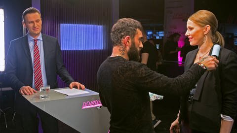 Drei Personen in einem TV-Studio, links ein Mann im Anzug hinter einem Pult, recht ein Mann mit Tattoo am Nacken, der einer blonden Frau mit einem Fusselroller über den Blazer streift. Sie blickt zu dem Mann hinter dem Pult und lächelt. (Foto: IMAGO, IMAGO / Russian Look)