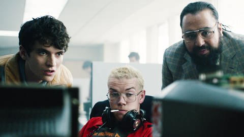 Drei Männer im Neunziger-Jahre-Look blicken in der Folge „Bandersnatch“ der Netflix-Serie „Black Mirror“ in einen Bildschirm: Fionn Whitehead, Will Poulter, Asim Chaudhry.