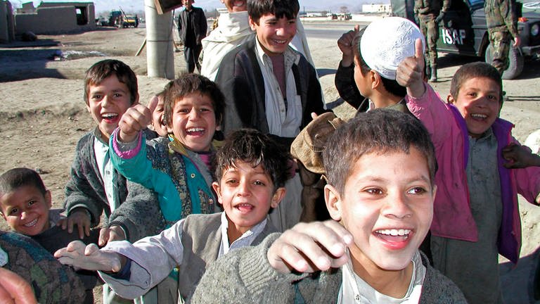 Eine Gruppe afghanischer Jungen die auf die fotografierende Person zulaufen und in die Kamera lachen und winken. Sie tragen unsortierte Kleidung, Wolljäckchen zu traditionellen längeren Gewändern. Die Kinder sind von ungefähr vier Jahren bis Teenageralter alt. Die Teenager halten sich eher im Hintergrund. Im Hintergrund sind auch mehrere Bundeswehrsoldaten zu sehen, die vor einem Panzerfahrzeug stehen.