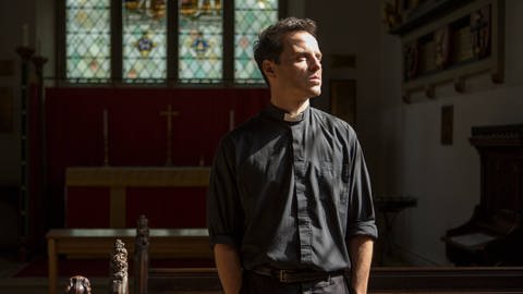 Andrew Scott als "heißer Priester" aus "Fleabag". Der Priester steht in seiner Kirche inmitten der Kirchenbänke.