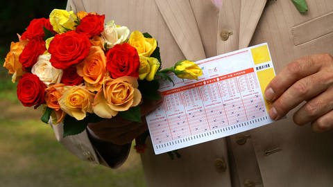 Mann mit Blumenstrauß und Lottoschein (Foto: IMAGO, IMAGO / McPHOTO)