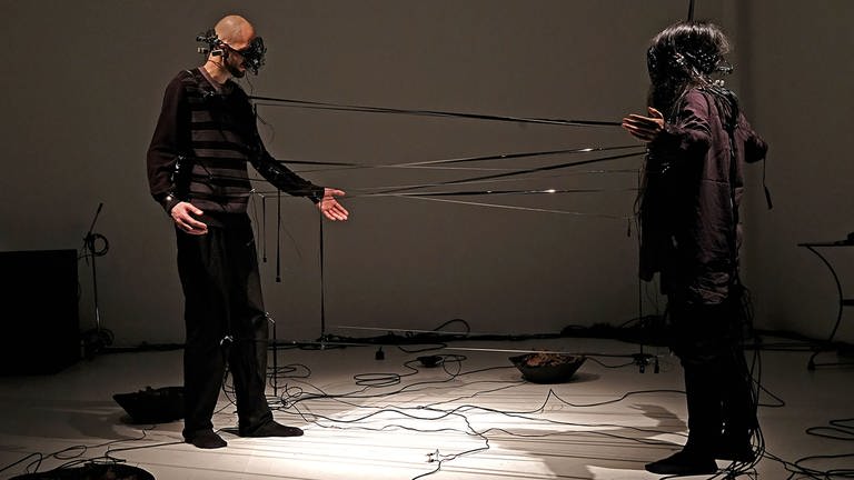 zwei Personen mit Apparaten vor den Augen, verbunden durch Kabel